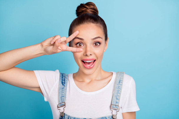 Close up photo of cheerful millennial making v-signs near face screaming wearing white t-shirt denim jeans overalls isolated over blue background - Photo, Image