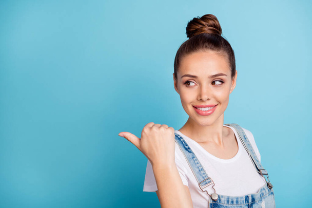 Portrait of charming lady showing at copy space by her forefinger looking wearing white t-shirt denim jeans overalls isolated over blue background - Zdjęcie, obraz