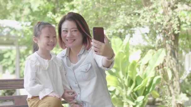 Азиатская веселая мать с дочерью, сидящей в саду и дающей селфи фото на смартфоне
. - Кадры, видео