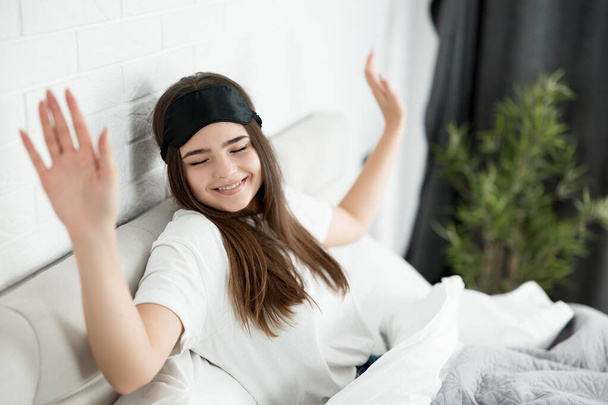 jeune femme attrayante avec masque de sommeil sur la tête assise dans le lit dans la chambre lumineuse bâillant le matin
 - Photo, image