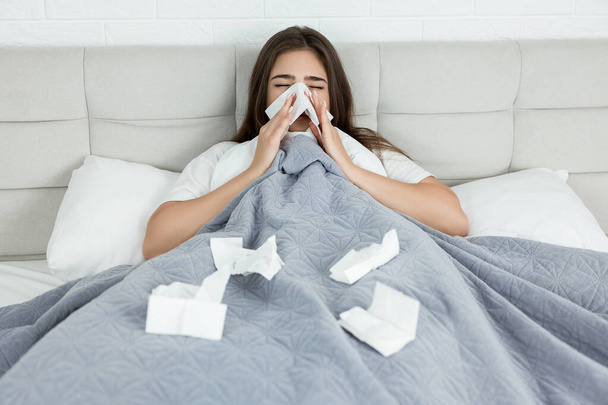 young sick woman sitting in bed bowing out her running nose with paper towel early in the morning - Fotoğraf, Görsel
