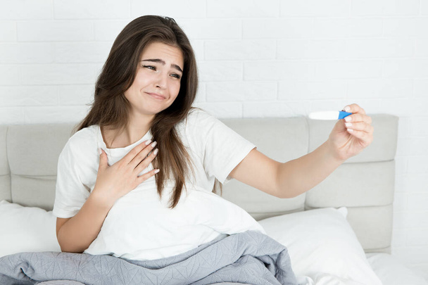 young attractive smiling woman sitting in bed holding pregnancy test in her hands early in the morning looking sentimental and happy - Foto, Imagen
