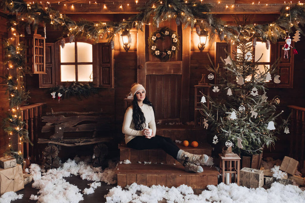 Brunette in hat with hot drink on wooden porch under snowfall. - Photo, image