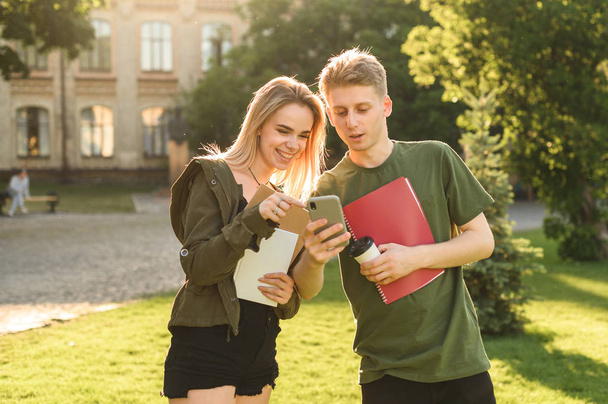 Deux étudiants heureux vérifiant le contenu du téléphone intelligent debout dans le parc de l'université tenant des livres et des cahiers. Quelques étudiants joyeux sur le terrain de l'université regardant le smartphone
. - Photo, image