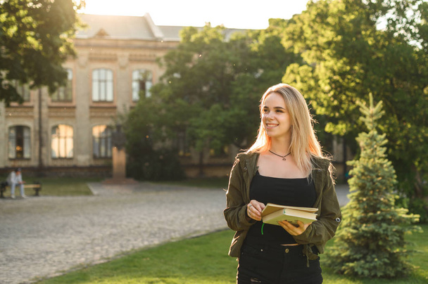 Piękna studentka z szerokim uśmiechem trzyma notebooki i patrząc na bok w ogrodzie kolegium. Uśmiechnięta atrakcyjna śliczna uczennica z książkami i podręczników w parku w pobliżu Uniwersytetu. - Zdjęcie, obraz