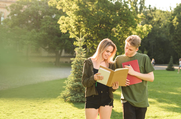大学キャンパスの近くの公園で宿題をチェックする2人の陽気な学生。学校の公園で本を読む肯定的な学生のカップル。不注意な成功した学生は、試験の結果を見て幸せ. - 写真・画像