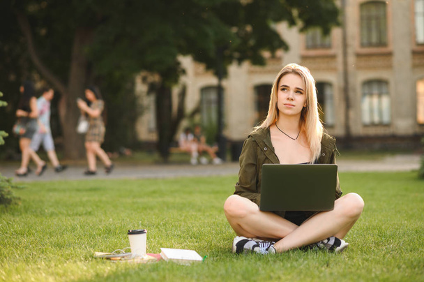 Zamyšlená studentka sní o tom, jak se dívá nahoru, zatímco pracuje na přenosném počítači, který sedí na univerzitě na zeleném trávníku a na pozadí univerzity. Kavkazská dívka s počítačem, knihami a kávou. - Fotografie, Obrázek