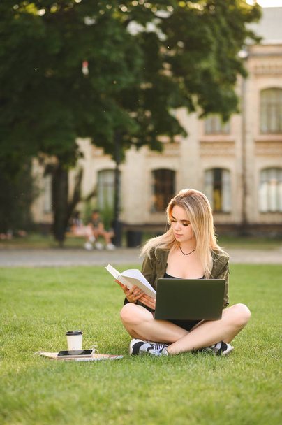 Pozytywna studentka siedzi Cross-legged na trawie z laptopem i książek robi pracę domową, przygotowując się do zajęć. Skoncentrowana dziewczyna kolegium siedzi na trawniku, czytanie książki uważnie. Strzał pionowy. - Zdjęcie, obraz
