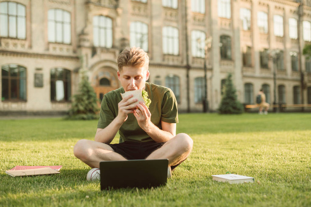 Πεινασμένος φοιτητής κάθεται στο γκαζόν στο πίσω μέρος του κτιρίου του Πανεπιστημίου, τρώγοντας ένα σάντουιτς και κοιτάζοντας την οθόνη ενός φορητού υπολογιστή, τα βιβλία είναι γρασίδι. Φοιτητής που σπουδάζει για διάλειμμα και φαγητό. - Φωτογραφία, εικόνα