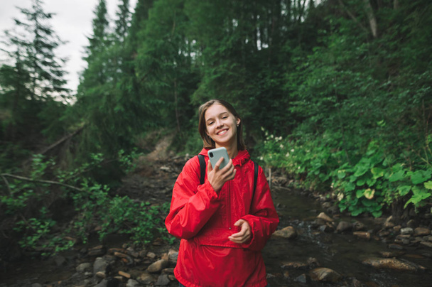 スマートフォンを手に森の中で笑みを浮かべてハイカーの女の子の肖像画、赤いレインコートを着て、渓流や野生植物の背景に立っている。ハイキング中の幸せな女の子はスマートフォンを使用しています. - 写真・画像
