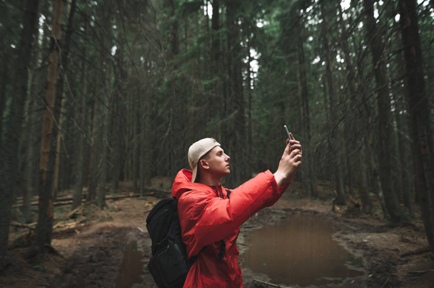 Jongeman in een rood jasje staat in het bos tijdens een wandeling en maakt een foto op een smartphone camera.Portret van een wandelaar in het bos met telefoon in de hand op de achtergrond van een bergpad met plas. - Foto, afbeelding