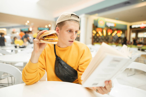 Portré egy koncentrált fiatalember elegáns könnyű ruházati, ül egy kávézóban egy szendvics a kezében, és gondosan olvassa el a könyv.Diák tanul és ételek egy gyorsétterem. - Fotó, kép