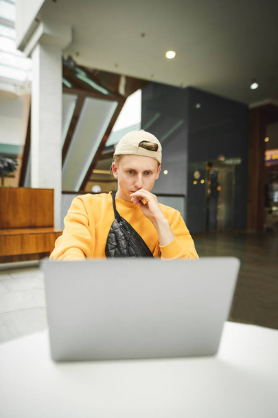 Όμορφος νεαρός άνδρας σε ένα καπέλο και ένα κίτρινο κρίνο λειτουργεί με έναν υπολογιστή, επικεντρώνεται στην εξέταση της οθόνης laptop ενός laptop. Κάθετη πορτρέτο ανεξάρτητος άνθρωπος εργάζεται ως ένα φορητό υπολογιστή σε ένα εμπορικό κέντρο. - Φωτογραφία, εικόνα