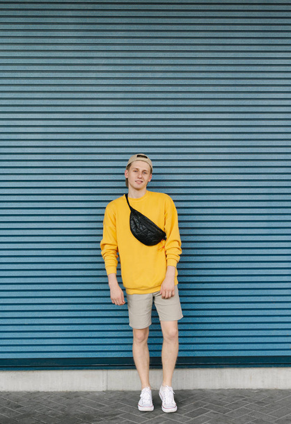 Retrato de um jovem em altura total, vestindo uma rua roupas casuais e uma mochila fanny, olhando para a câmera e sorrindo. Cara em estilo de rua posa contra o fundo de uma parede azul
. - Foto, Imagem