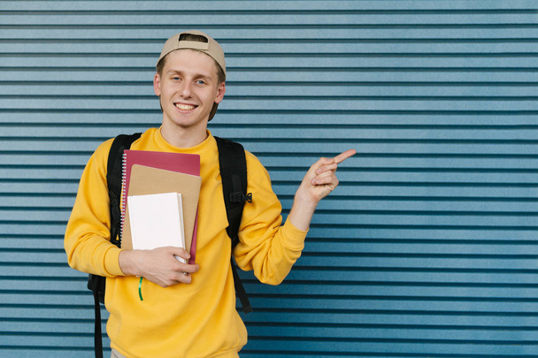 Glimlachende stijlvolle student met boeken en notitieboekjes in zijn handen staat tegen de achtergrond van een blauwe muur en toont zijn vinger opzij naar een lege ruimte, met casual kleding en een rugzak.Copyspace - Foto, afbeelding