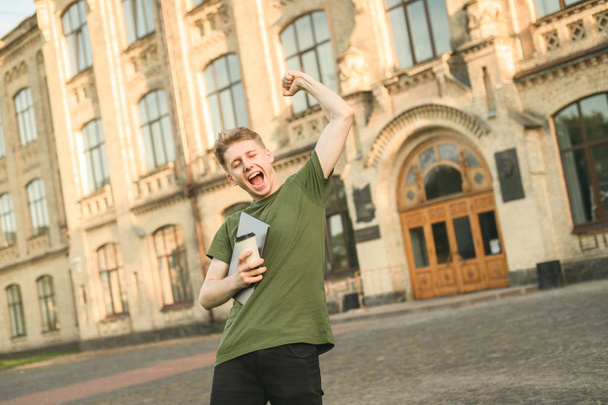 Happy cheerful excited male student celebrating his victory with raised hands in front of college campus. The main task completed, exams over, reaching success, winner student with laptop and coffee. - Photo, Image