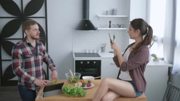 cheerful girl sitting on table and taking pictures funny guy on cell phone while cooking useful meal for brunch - Footage, Video
