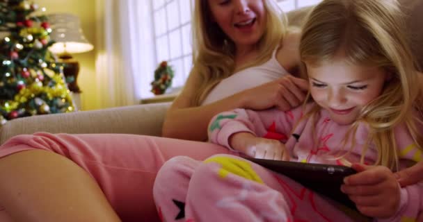 Front view close up of a happy young Caucasian mother sitting on the sofa with her young daughter using a tablet in their sitting room at Christmas time, the mother tickling her daughter - Metraje, vídeo