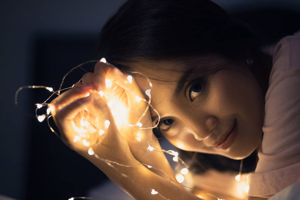 Une jeune femme couchée et jouant avec des lumières scintillantes ou une lumière de fée sur un lit blanc Dans une pièce avec une faible lumière jaune
. - Photo, image