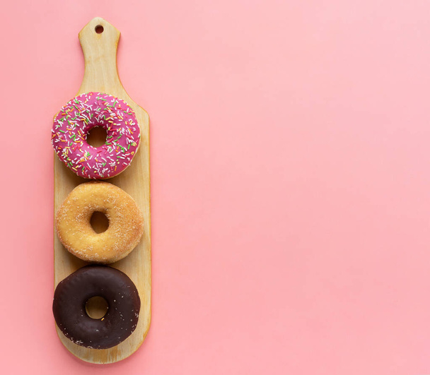Τρία διαφορετικά ντόνατς, ντόνατ σοκολάτας, ροζ κερασάκι και χωρίς γλάσο σε ξύλινο ξύλο κοπής σε ροζ φόντο - Φωτογραφία, εικόνα