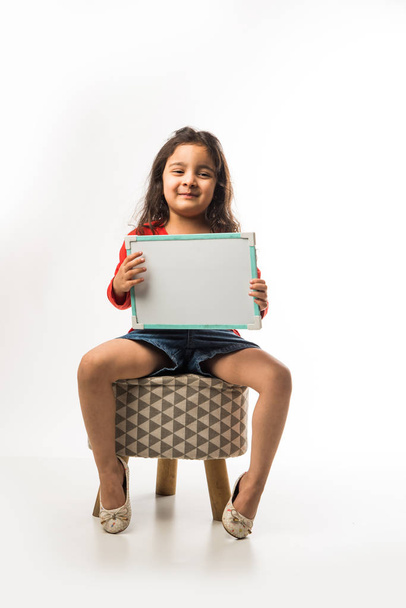 small Indian girl writing on slate with marker pen, while sitting on stool over white background - Photo, image