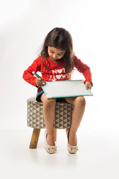small Indian girl writing on slate with marker pen, while sitting on stool over white background - Фото, изображение