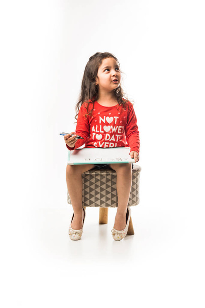 small Indian girl writing on slate with marker pen, while sitting on stool over white background - Foto, Imagem