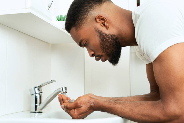 Черный парень моет лицо в ванной комнате, вид сбоку
 - Фото, изображение