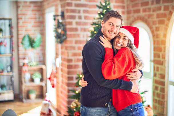 Young beautiful couple smiling happy and confident. Standing and hugging around christmas tree at home - Zdjęcie, obraz