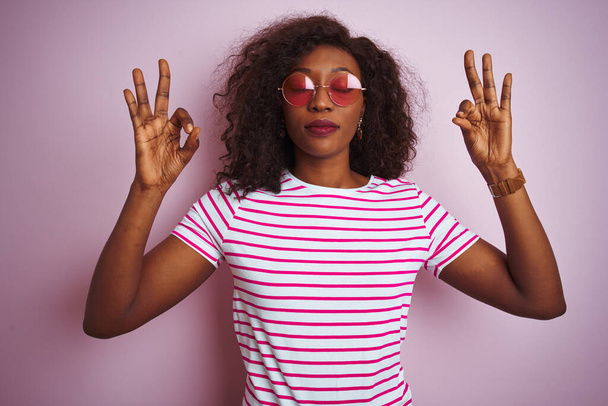 Jonge Afro-Amerikaanse vrouw draagt t-shirt en zonnebril over geïsoleerde roze achtergrond ontspannen en glimlachen met de ogen dicht doen meditatie gebaar met de vingers. Yoga-concept. - Foto, afbeelding