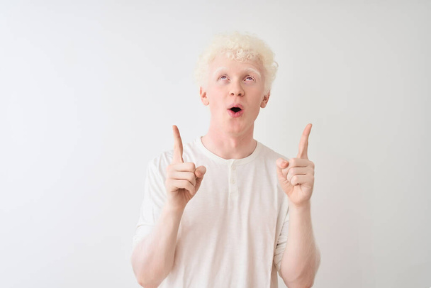 Young albino blond man wearing casual t-shirt standing over isolated white background amazed and surprised looking up and pointing with fingers and raised arms. - Photo, Image