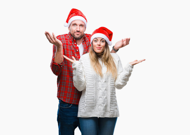 Молодая пара влюблена в рождественскую шляпу на изолированном фоне невежественное и запутанное выражение лица с поднятыми руками и руками. Концепция сомнений
. - Фото, изображение