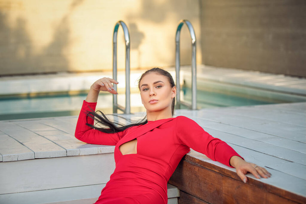 Fashion brinette vrouw in lange rode jurk liggend bij het zwembad - Foto, afbeelding