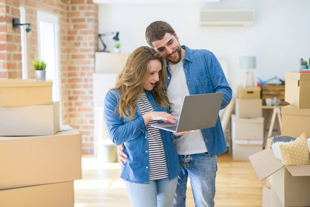Молодая пара, использующая ноутбук, стоящий в комнате вокруг картонных коробок, счастлива переехать в новую квартиру
 - Фото, изображение