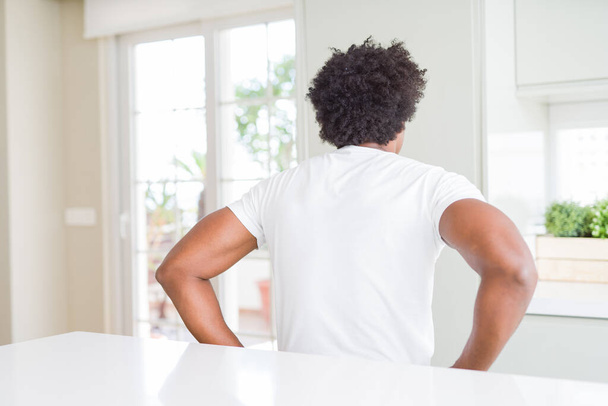 Young african american man wearing casual white t-shirt sitting at home standing backwards looking away with arms on body - Foto, afbeelding