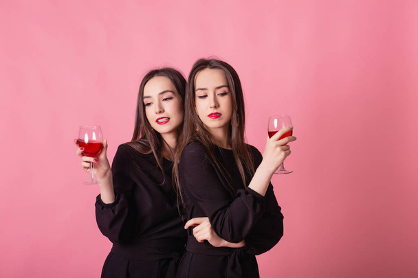 Two beautiful girls twins celebrate corporate party and drinking wine. Girls posing on pink background, bright makeup, red lips, wearing black stylish evening outfits. - Photo, image
