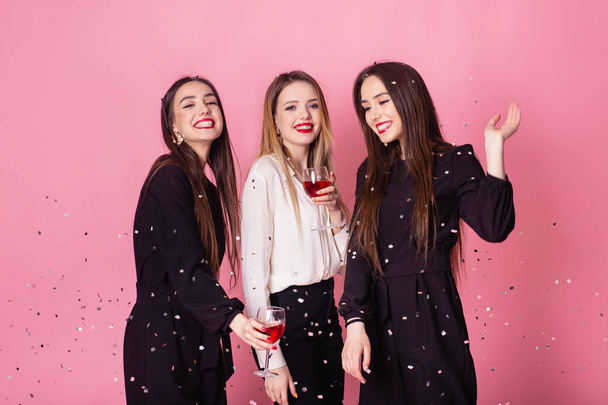 Trzy kobiety świętują Nowy Rok, śmiejąc się pod latającym konfetti i pijąc wino. Dziewczyny pozowanie i uśmiech na różowym tle, pogodne emocje, jasny makijaż, wewnątrz strony, noszenie czarno-białe stylowe stroje wieczorowe. - Zdjęcie, obraz