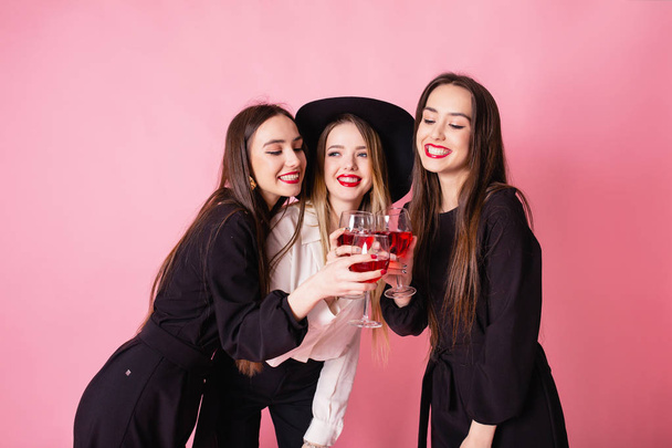 Tres hermosas chicas celebran la fiesta corporativa y beben vino. Chicas posando sobre fondo rosa, maquillaje brillante, labios rojos, con trajes de noche elegantes en blanco y negro
. - Foto, Imagen