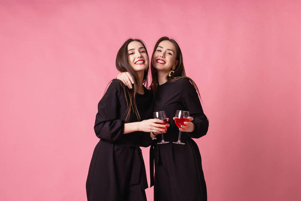 İki güzel kız ikiz şirket yılbaşı partisini kutluyor, şarap içip sohbet ediyorlar. Pembe arka planda poz veren kızlar, parlak makyaj, kırmızı dudaklar, siyah şık gece kıyafetleri giyen kızlar.. - Fotoğraf, Görsel