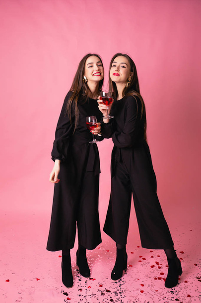 Two beautiful girls twins celebrate corporate New Year party, drinking wine and talking. Girls posing on pink background, bright makeup, red lips, wearing black stylish evening outfits. - Photo, Image