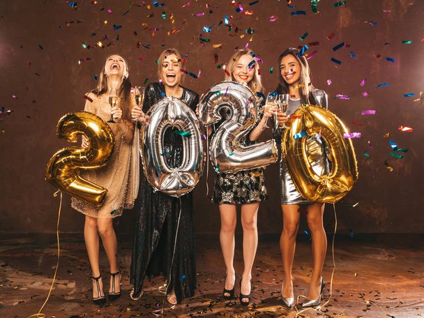 Belles femmes célébrant le Nouvel An. Filles magnifiques heureux dans des robes de soirée sexy élégantes tenant or et argent 2020 ballons, s'amuser à la fête du Nouvel An. Arrangeant et buvant des flûtes à champagne
 - Photo, image