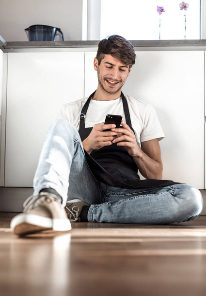young man talking on mobile phone sitting on kitchen floor - Zdjęcie, obraz