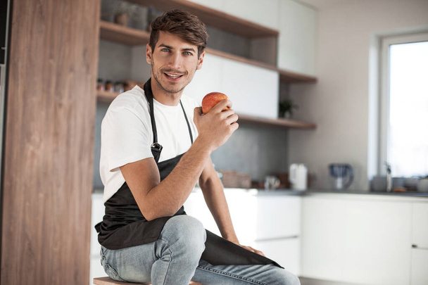 attractive man with an apple sitting in a home kitchen - Photo, image