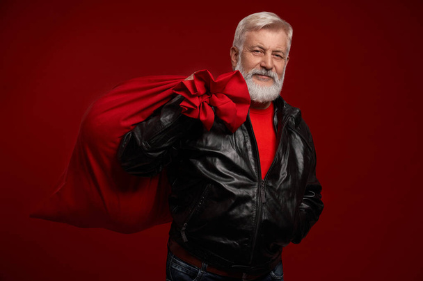 Mature man wearing leather jacket holding red Santa bag - Фото, зображення