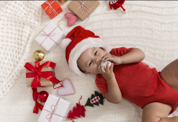 Симпатичный малыш, кусающий рождественский шар, лежит среди коробок, вид сверху
 - Фото, изображение