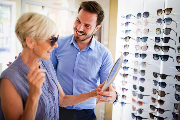 Health care, eyesight and vision concept. Happy senior woman choosing glasses at optics store - Фото, изображение