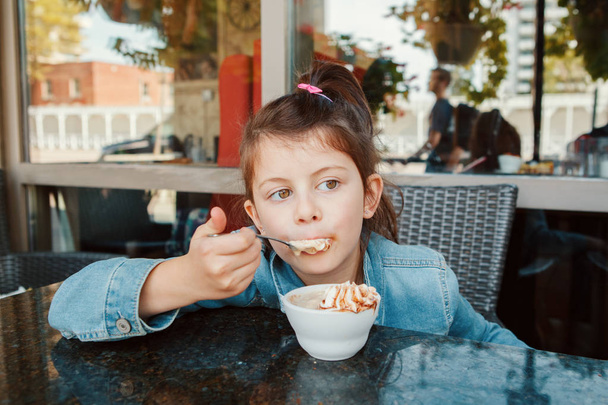 Funny Caucasian preschool girl eating sweet dessert with spoon in cafe. Child kid having fun in restaurant patio enjoying food drink. Happy authentic childhood lifestyle.  - 写真・画像