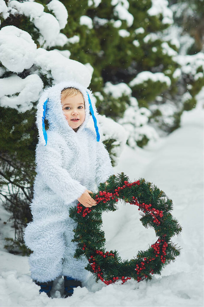 little boy dressed in rabbit costume holding  christmas wreath in snowy winter park, new year concept    - Photo, Image