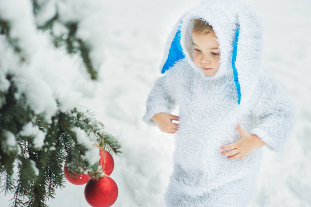 little boy dressed in rabbit costume standing nera decorative Christmas tree in snowy winter park, new year  concept   - 写真・画像