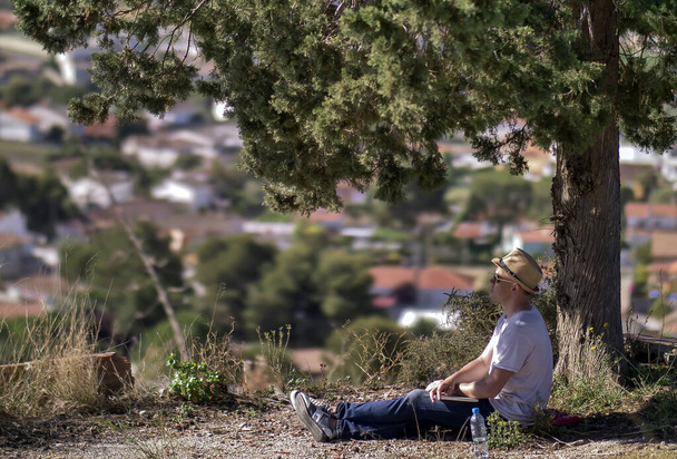Młody człowiek z laptopem i kapeluszem siedzi pod drzewem na rozmazanym tle miasta - Zdjęcie, obraz